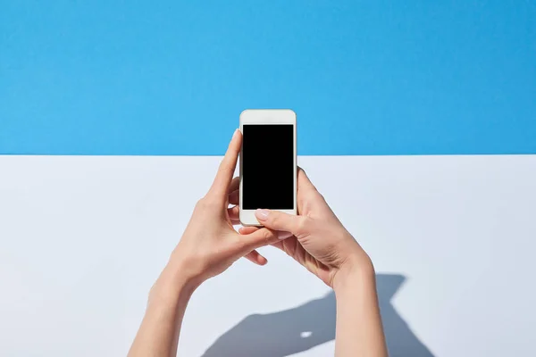 裁剪的妇女的看法使用智能手机与空白屏幕上的白色办公桌和蓝色背景 — 图库照片
