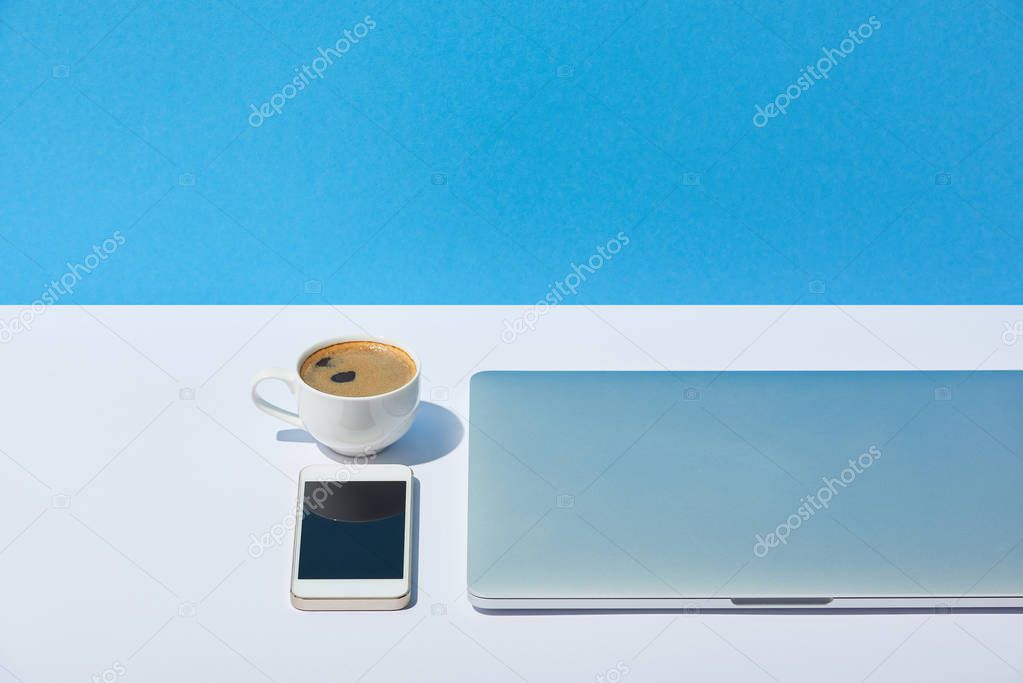 laptop, coffee cup and smartphone on white desk and blue background