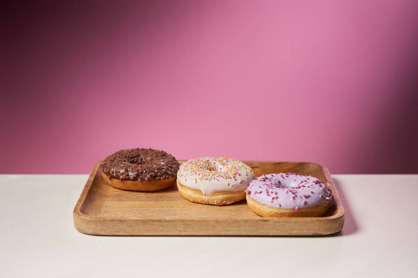 tasty donuts with icing on wooden cutting board on pink background 