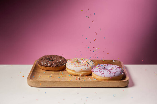 tasty donuts with falling sprinkles on wooden cutting board on pink background 