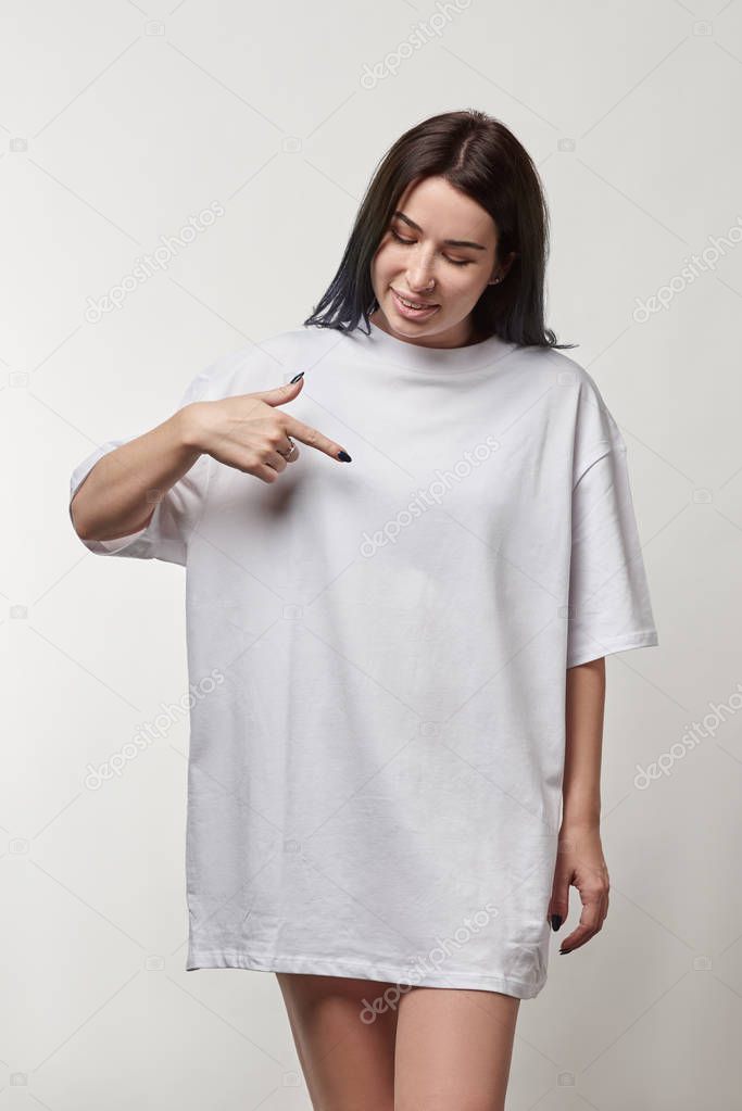 beautiful young woman pointing with finger at white oversize t-shirt with copy space isolated on grey