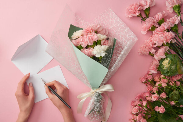 обрезанный снимок женщины, пишущей на поздравительной открытке и красивый розовый букет цветов, изолированный на розовый

