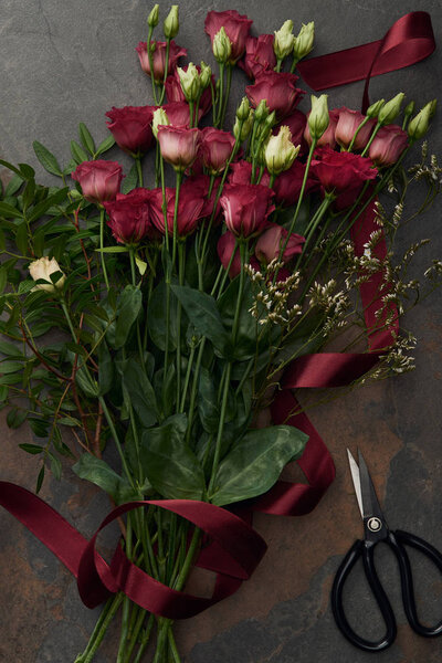 top view of beautiful red eustoma flowers with ribbon and scissors on dark surface   