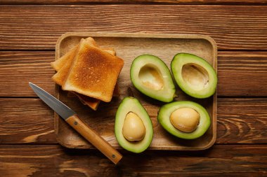 top view of cut avocados, grilled crispy toasts and knife on wooden background clipart