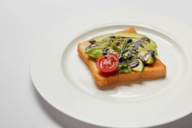 selective focus of toast with avocado and cherry tomato on white plate and grey background clipart