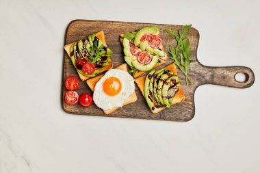top view of wooden cutting board with toasts, scrambled egg and cherry tomatoes on marble surface clipart