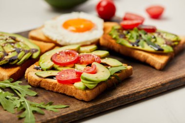 selective focus of cutting board with toasts, scrambled egg, cherry tomatoes and avocado on white background