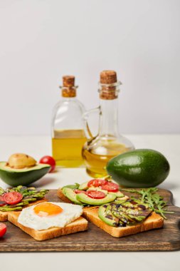 Ahşap kesme tahtası tost, çırpılmış yumurta, domates, avokado ve gri arka plan üzerinde yağ şişeleri ile seçici odak