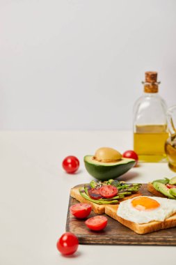 selective focus of wooden cutting board with toasts, scrambled egg, cherry tomatoes, avocados and oil bottles on grey background clipart