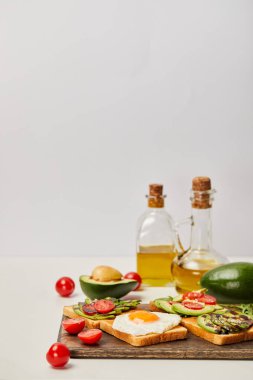 Ahşap kesme tahtası tost, çırpılmış yumurta, domates, avokado ve gri arka plan üzerinde yağ şişeleri ile seçici odak