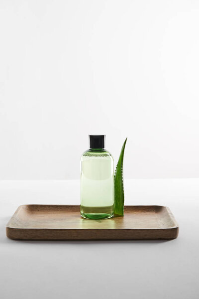 transparent cosmetic bottle and aloe vera leaf on wooden tray on white surface