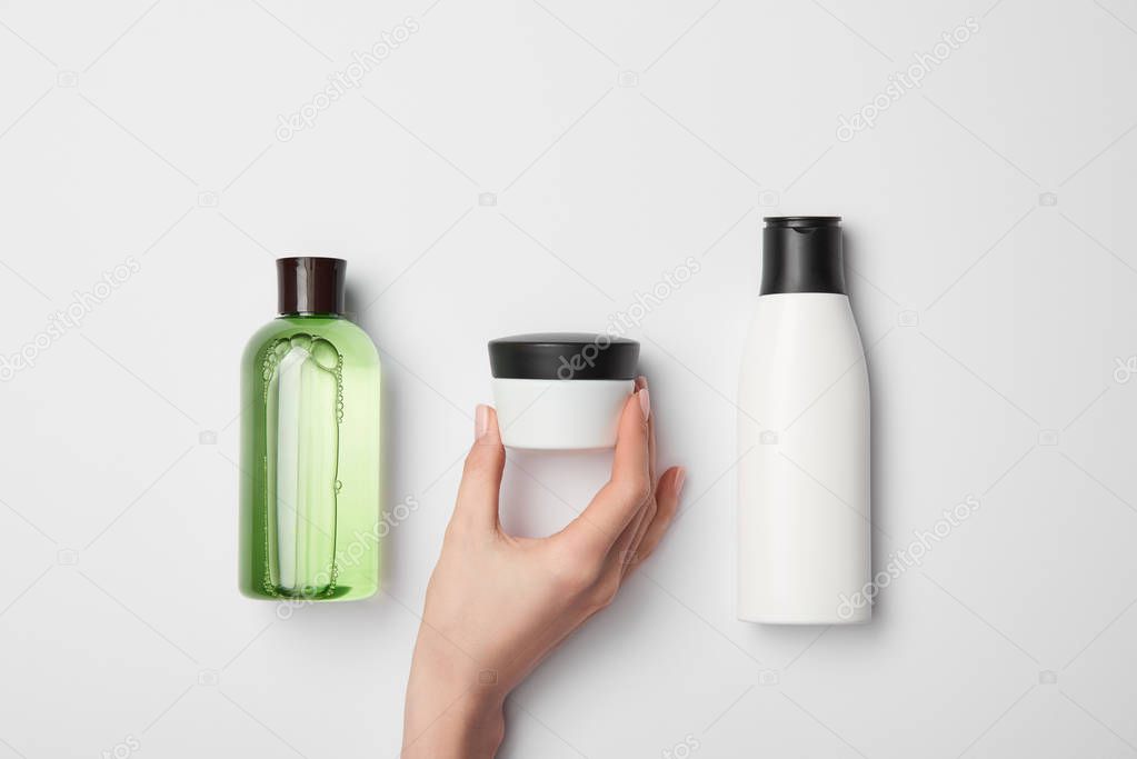 Top view of female hand near cream container with different cosmetic bottles on white background