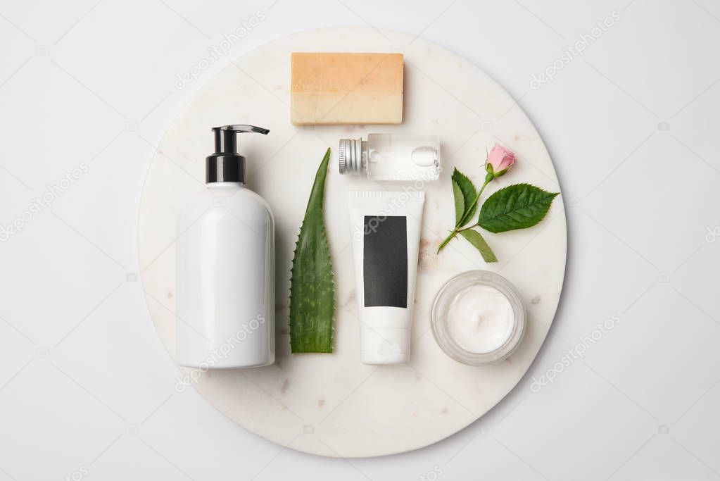 Top view of different cosmetic containers, soap, aloe vera leaf and rose flower on white round surface