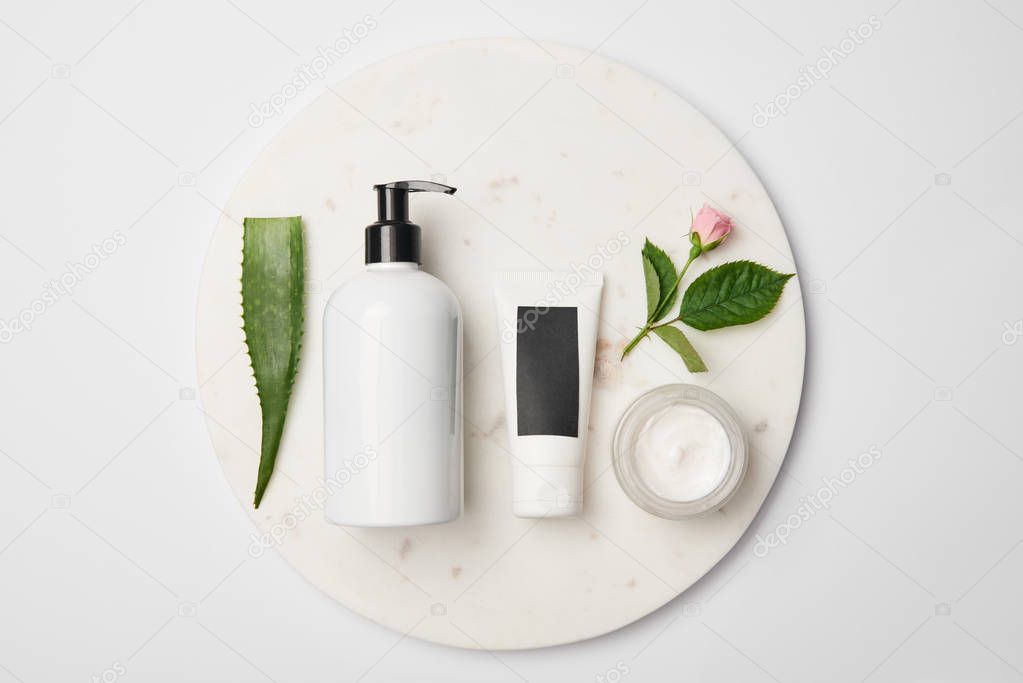 Top view of different cosmetic containers, aloe vera leaf and rose flower on white round surface