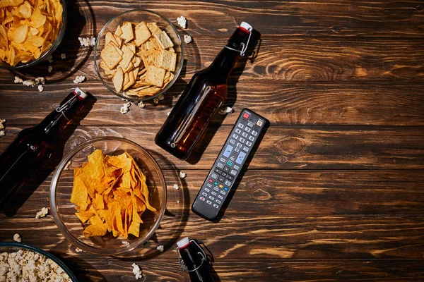 top view of snacks near bottles with beer and remote control on wooden table