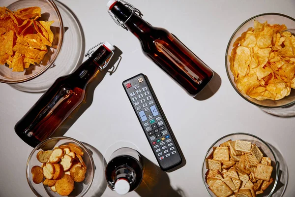 top view of brown bottles with beer near snacks in bowls and remote control on white background