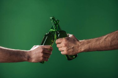 partial view of men clinking beer bottles isolated on green, st patrick day concept clipart