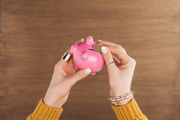 cropped view of woman puting in piggy bank coin on wooden background