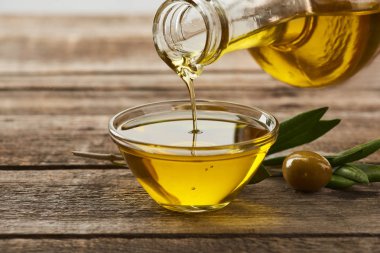 pouring oil from bottle into glass bowl, olive tree leaves and olive on wooden surface clipart