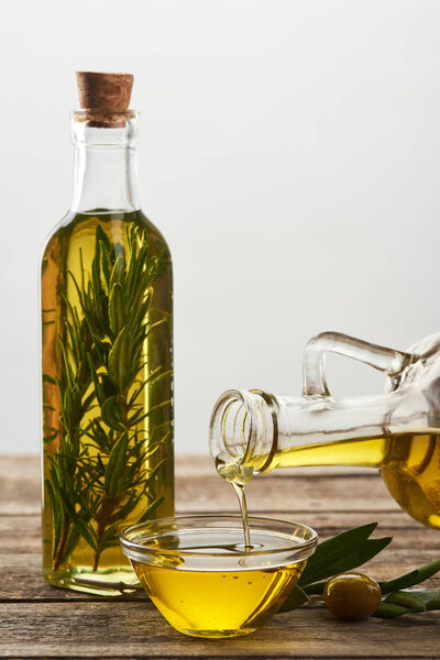 pouring olive oil from bottle into glass bowl, bottle of oil flavored with rosemary, olive tree leaves and olives on wooden surface
