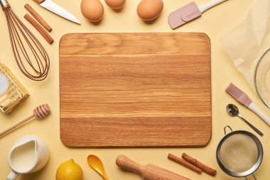 top view of empty wooden chopping board with cooking utensils and ingredients on yellow background  clipart