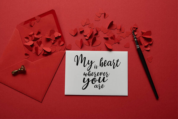 top view of envelope with "my heart is wherever you are" lettering , paper cut hearts and pen on red background