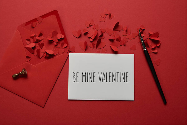 top view of envelope with "be mine valentine" lettering , paper cut hearts and pen on red background