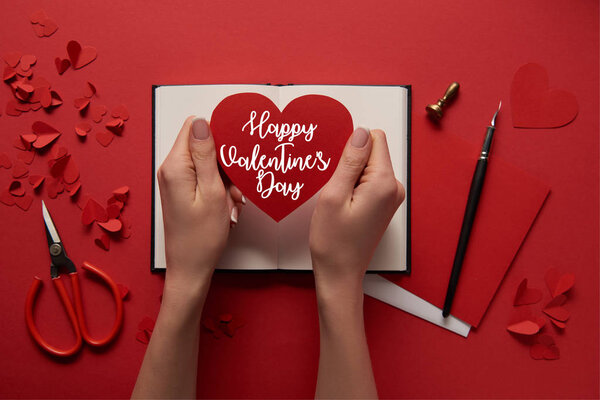 cropped view of woman holding paper cut heart sign with "Happy valentines day" lettering on red background