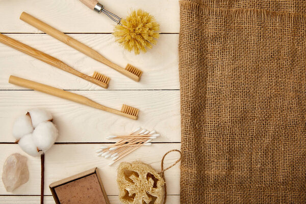 flat lay of different hygiene and care items, and piece of sackcloth on white wooden surface, zero waste concept