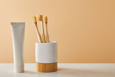 bamboo toothbrushes in holder and toothpaste in tube on white table and beige background clipart