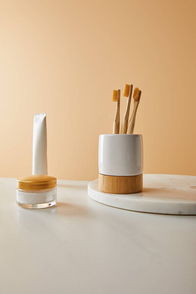 toothpaste in tube, cosmetic cream and holder with bamboo toothbrushes on white marble surface and beige background
