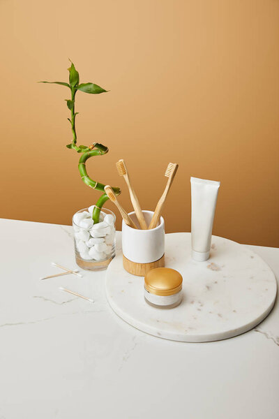 bamboo toothbrushes in holder, cosmetic cream and toothpaste in tube on round board near green bamboo stem in vase on beige background