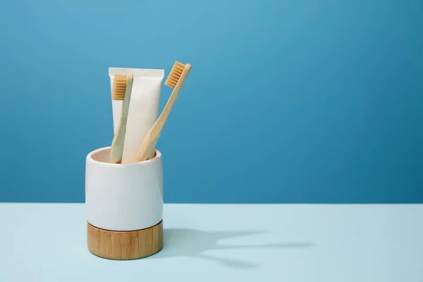 holder with toothpaste in tube and bamboo toothbrushes on table and blue background