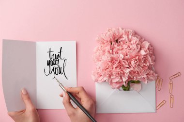 cropped view of woman holding greeting card with trust your soul lettering and pen near carnation flowers in envelope on pink background clipart