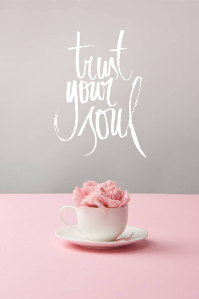 pink carnation flowers in white cup on saucer on grey background with trust your soul lettering