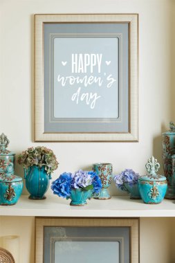 picture frame with happy womens day lettering and turquoise ceramic ornate vintage vases with flowers on shelf clipart