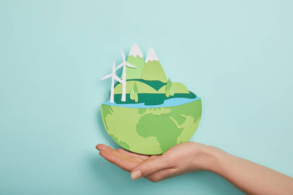top view of woman holding paper cut planet with renewable energy sources on turquoise background, earth day concept