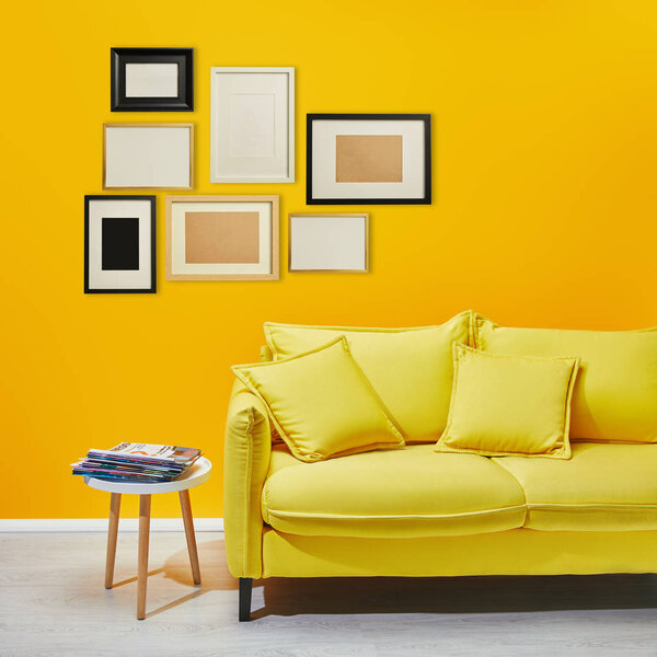 coffee table standing near modern yellow sofa near decorative frames hanging on wall