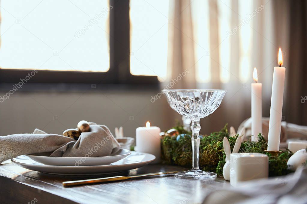 selective focus of quail eggs on napkin and plates near green moss, burning candles and crystal glass on wooden table at home