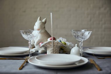 Beyaz tabak, çatal bıçak takımı, kristal gözlük, dekoratif bunnie ve sepet ile evde masada boyalı yumurta