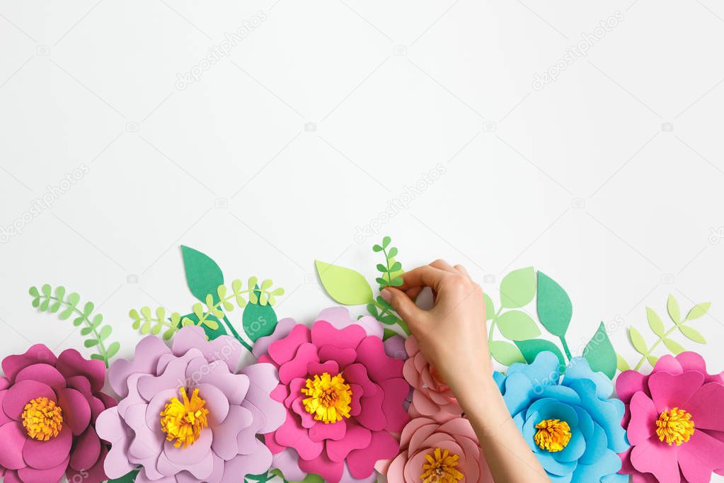 cropped view of woman putting green plant near multicolored paper flowers on grey background