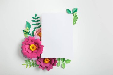 top view of white blank card near pink paper flowers and green leaves on grey background clipart
