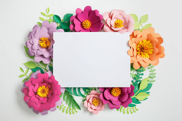 top view of white blank card on multicolored paper flowers with green leaves on grey background