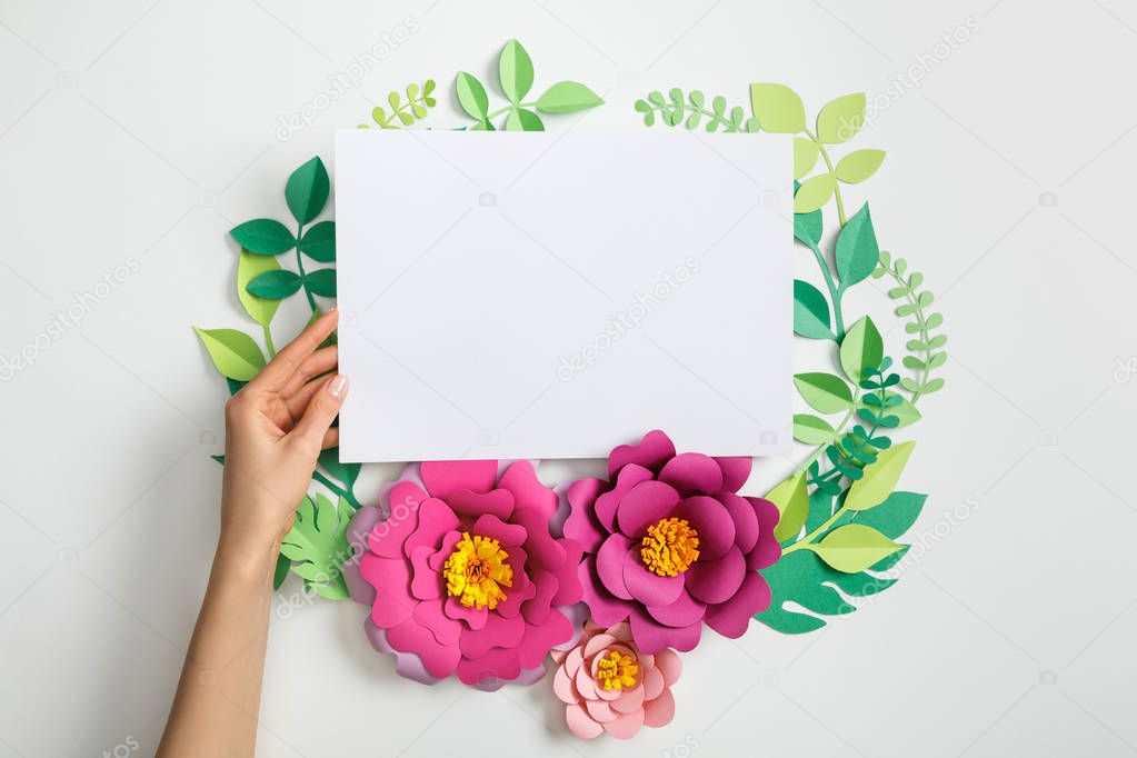 cropped view of woman putting white blank card near pink paper flowers and leaves on grey background