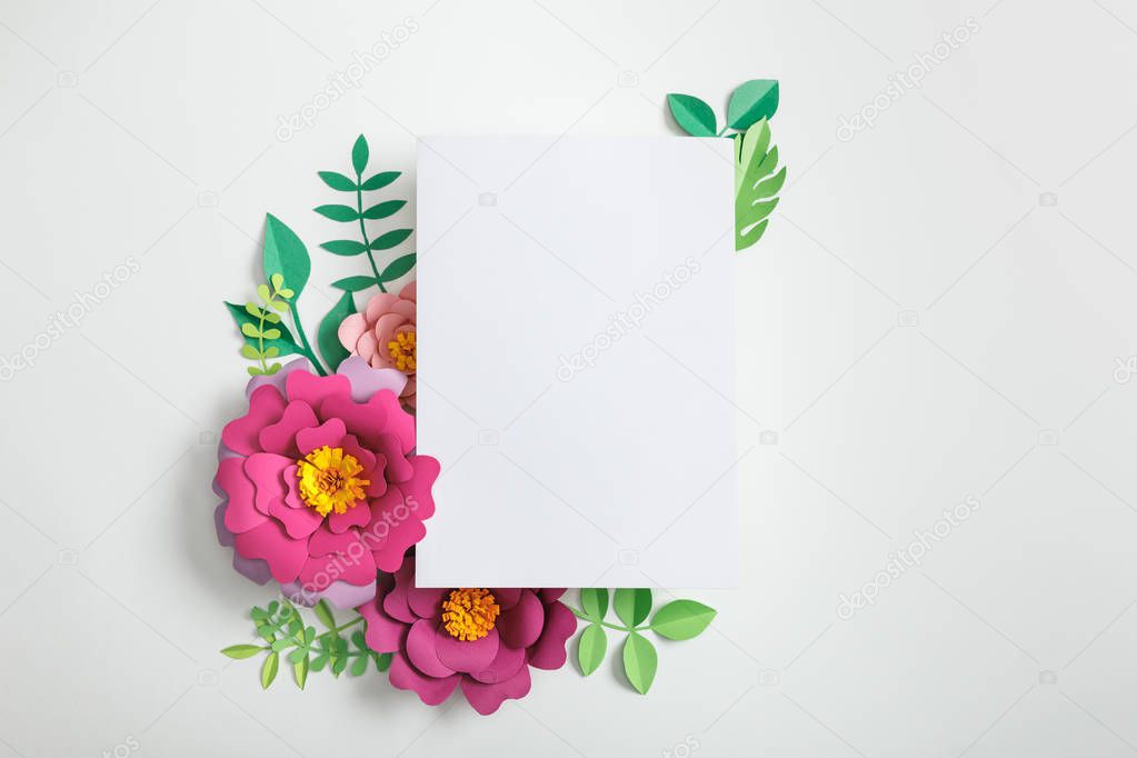 top view of white blank card near pink paper flowers and green leaves on grey background