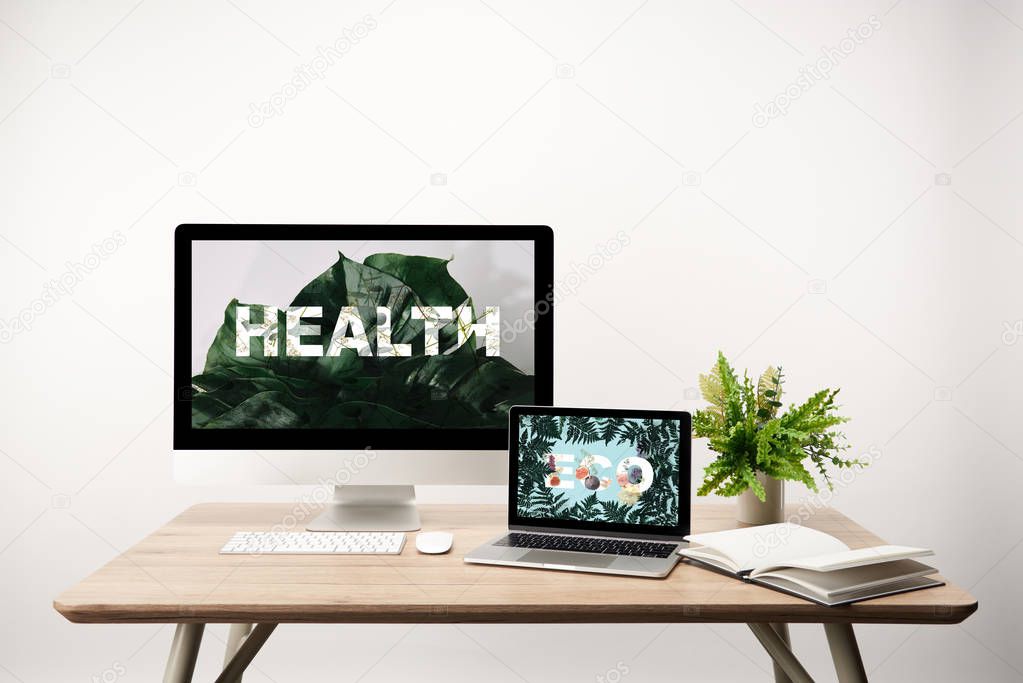 computer with health illustration and monstera leaves on monitor and laptop with eco lettering and fern leaves on screen on wooden table