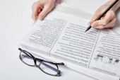 cropped view of woman holding pencil near newspaper and glasses on white 