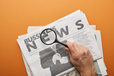 man holding magnifying glass and zooming news lettering while reading  business newspapers on orange  clipart