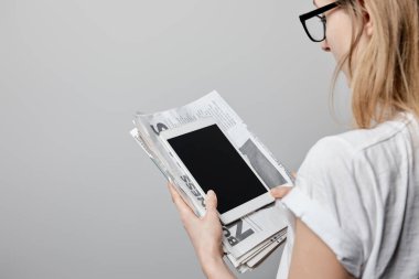 cropped view of woman in glasses holding digital tablet with blank screen and newspapers isolated on grey  clipart