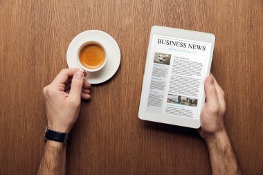 cropped view of man holding digital tablet with business news on screen and cup of coffee clipart